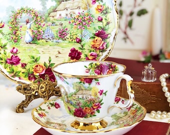 May include: A white teacup and saucer with a floral pattern featuring a cottage and roses. The teacup and saucer have gold trim. The teacup has a handle. The teacup and saucer are on top of a red book with the title "Adventures of Odysseus".