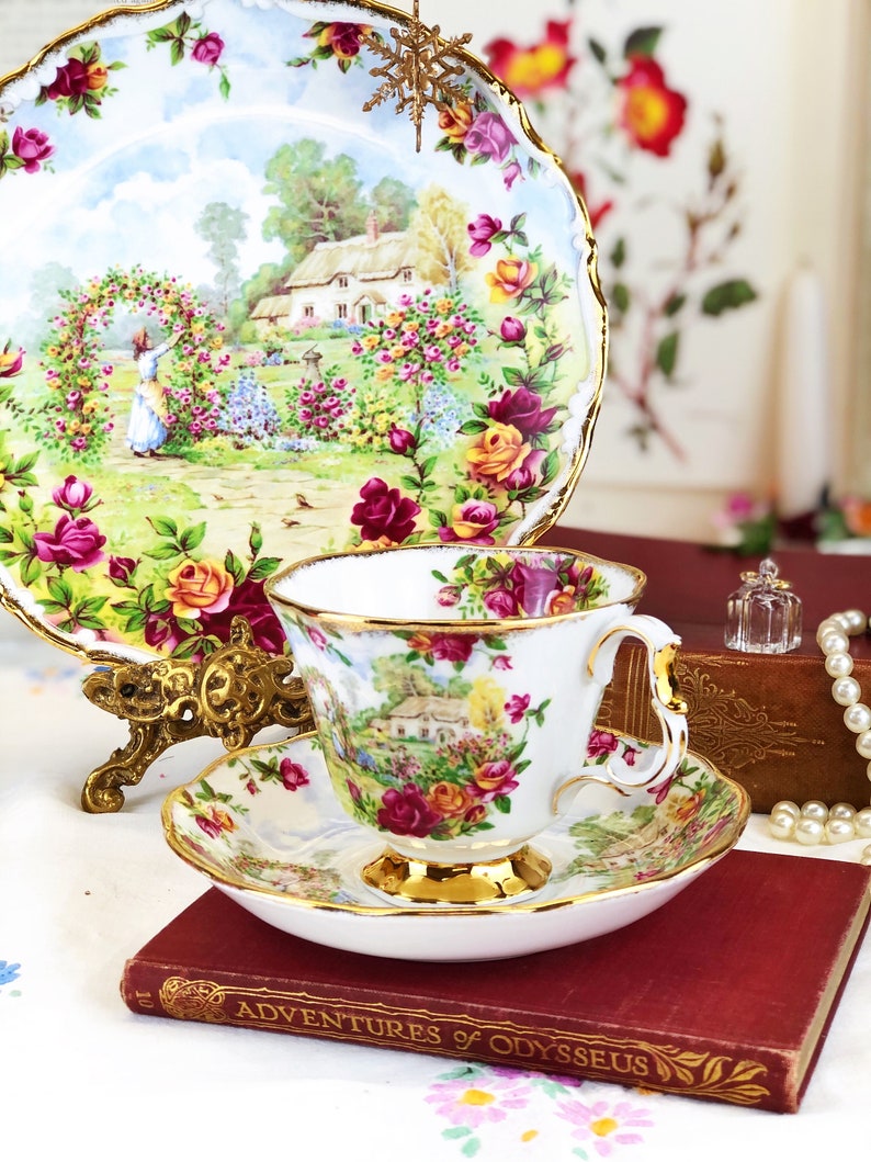 K&ouml;nnte beinhalten: Eine wei&szlig;e Teetasse und Untertasse mit einem Blumenmuster, das ein H&auml;uschen und Rosen zeigt. Die Teetasse und Untertasse haben einen goldenen Rand. Die Teetasse hat einen Henkel. Die Teetasse und Untertasse stehen auf einem roten Buch mit dem Titel "Adventures of Odysseus".