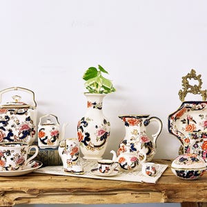Può includere: Una collezione di tazze da tè, teiera, lattiera, zuccheriera e vaso in stile vintage con motivi floreali in tonalità di blu, rosso e bianco. Il set è disposto su una superficie in legno con una pianta verde sullo sfondo.