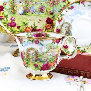 K&ouml;nnte beinhalten: Eine wei&szlig;e Teetasse und Untertasse mit einem Blumenmuster, das ein H&auml;uschen und eine Frau in einem Garten zeigt. Die Teetasse hat einen goldenen Rand und einen goldenen Henkel. Die Untertasse hat einen gewellten Rand.