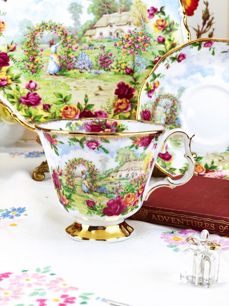 K&ouml;nnte beinhalten: Eine wei&szlig;e Teetasse und Untertasse mit einem Blumenmuster, das ein H&auml;uschen und eine Frau in einem Garten zeigt. Die Teetasse hat einen goldenen Rand und einen goldenen Henkel. Die Untertasse hat einen gewellten Rand.