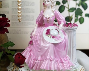 May include: A porcelain figurine of a woman in a pink dress sitting on a pedestal with roses. She is holding a white hat with a pink ribbon.