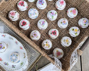 Könnte beinhalten: Ein Weidenkorb gefüllt mit kleinen, weißen Porzellan-Schmuckkästchen. Jede Schachtel ist mit einem anderen Blumendesign in Rosa-, Lila- und Gelbtönen verziert. Die Schachteln sind in Reihen angeordnet und bilden ein optisch ansprechendes Display.
