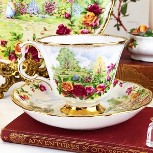 K&ouml;nnte beinhalten: Eine wei&szlig;e Teetasse und Untertasse mit einem Blumenmuster und goldenen Verzierungen. Die Teetasse zeigt eine gemalte Szene eines Gartens mit B&auml;umen und Blumen. Die Untertasse hat ein &auml;hnliches Blumenmuster und einen goldenen Rand.