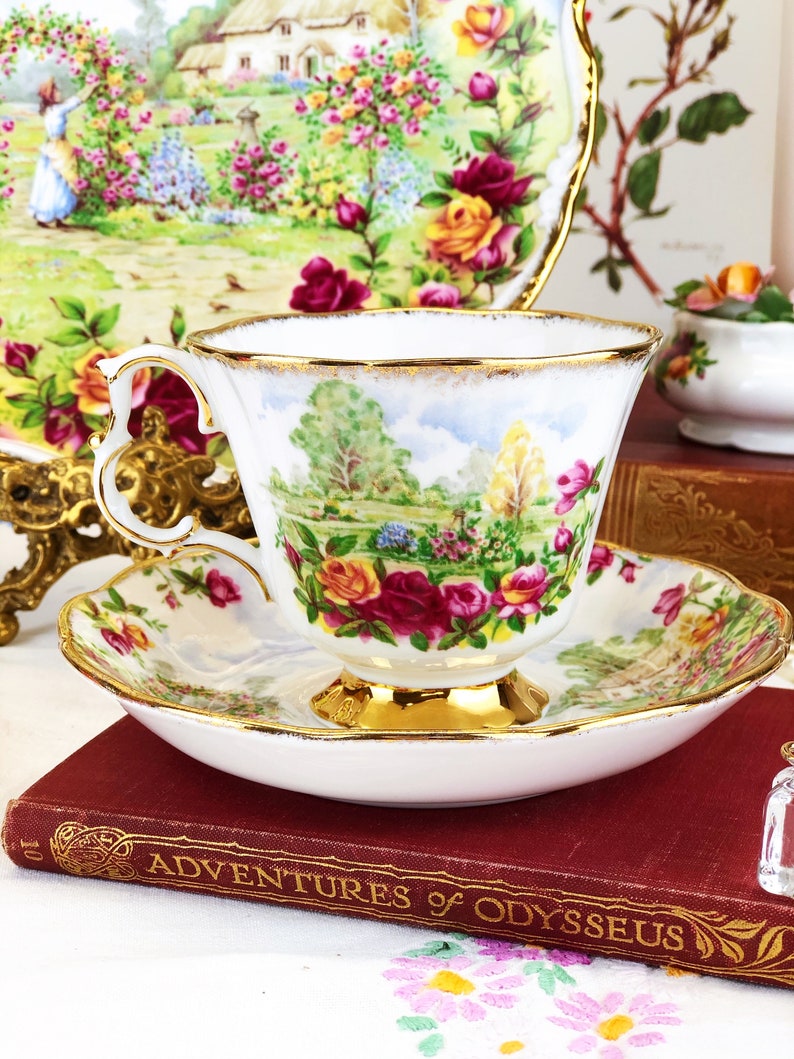 K&ouml;nnte beinhalten: Eine wei&szlig;e Teetasse und Untertasse mit einem Blumenmuster und goldenen Verzierungen. Die Teetasse zeigt eine gemalte Szene eines Gartens mit B&auml;umen und Blumen. Die Untertasse hat ein &auml;hnliches Blumenmuster und einen goldenen Rand.