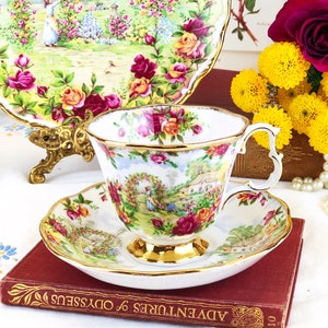 K&ouml;nnte beinhalten: Eine wei&szlig;e Teetasse und Untertasse mit einem Blumenmuster mit Rosen, einem H&auml;uschen und einer Gartenszene. Tasse und Untertasse haben einen goldenen Rand. Die Teetasse und Untertasse stehen auf einem roten Buch mit dem Titel "Adventures of Odysseus".