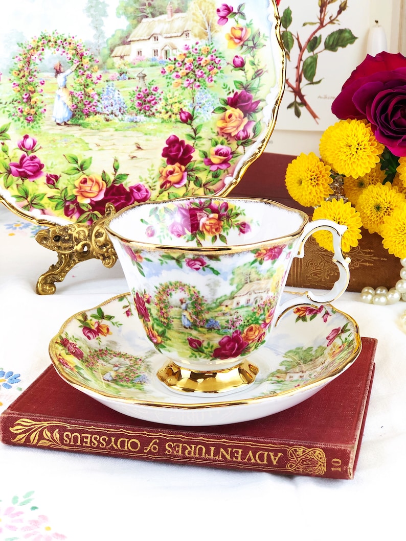 K&ouml;nnte beinhalten: Eine wei&szlig;e Teetasse und Untertasse mit einem Blumenmuster mit Rosen, einem H&auml;uschen und einer Gartenszene. Tasse und Untertasse haben einen goldenen Rand. Die Teetasse und Untertasse stehen auf einem roten Buch mit dem Titel "Adventures of Odysseus".