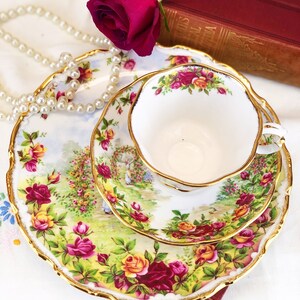 K&ouml;nnte beinhalten: Ein wei&szlig;es Tee-Set mit Untertasse und einem floralen Muster. Das Set verf&uuml;gt &uuml;ber einen goldenen Rand und ein zartes Blumenmuster mit roten, gelben und rosa Rosen. Das Set wird auf einer wei&szlig;en Tischdecke mit einer Perlenkette und einer roten Rose pr&auml;sentiert.