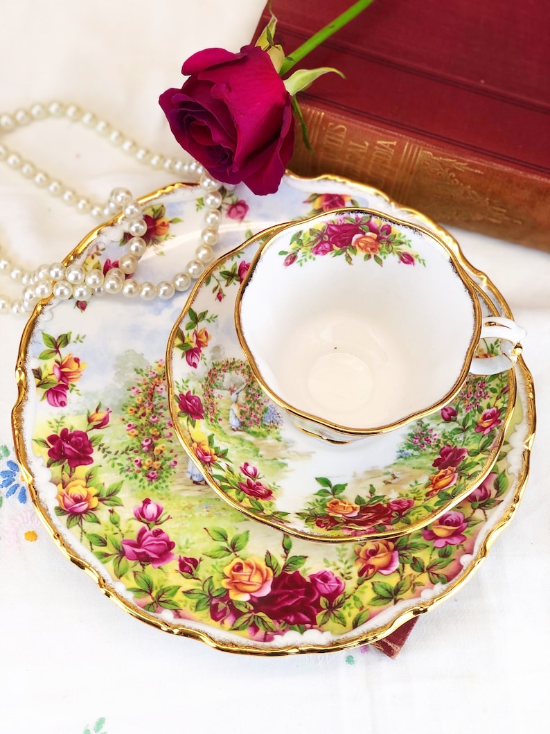K&ouml;nnte beinhalten: Ein wei&szlig;es Tee-Set mit Untertasse und einem floralen Muster. Das Set verf&uuml;gt &uuml;ber einen goldenen Rand und ein zartes Blumenmuster mit roten, gelben und rosa Rosen. Das Set wird auf einer wei&szlig;en Tischdecke mit einer Perlenkette und einer roten Rose pr&auml;sentiert.