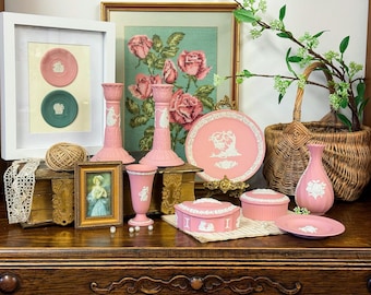Puede incluir: Una colección de objetos decorativos rosas y blancos, incluyendo candelabros, un jarrón, un plato y una caja de baratijas, dispuestos sobre una superficie de madera. Una obra de arte enmarcada y una cesta de mimbre también están presentes, creando una estética vintage.