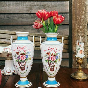 May include: Two white porcelain vases with blue and gold detailing, decorated with floral arrangements and red bows. Each vase has a handle and contains red tulips. The vases are on a dark wooden table.