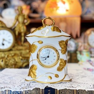 May include: White porcelain clock with gold accents and a leaf design. The clock face has Roman numerals and black hands. The clock is sitting on a book with the text "CASSEL HISTO OF ENGLA VOL.I."
