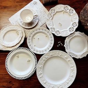 Puede incluir: Una colección de platos de cerámica blanca y una taza de té con intrincados diseños de encaje. Los platos varían en forma y tamaño, algunos con patrones florales en relieve y otros con delicados bordes calados. Algunas perlas falsas están dispersas.