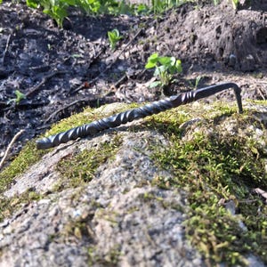 Puede incluir: Una herramienta de jardín de metal oscuro forjada a mano, con un mango retorcido y un extremo en forma de gancho. La herramienta descansa sobre una roca cubierta de musgo, con un fondo borroso de tierra, plantas y árboles. El diseño de la herramienta sugiere que es para cavar o desherbar.