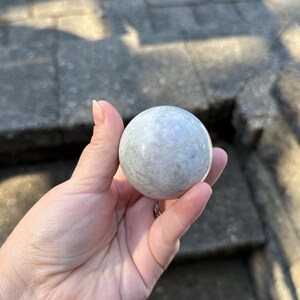 May include: A light gray sphere with subtle purple and green veining is held in a hand. The sphere is smooth and polished, reflecting the sunlight. The object is a decorative item, possibly a crystal or gemstone.