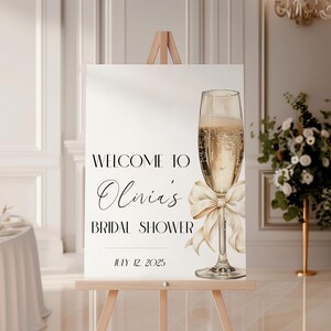 May include: Welcome to Olivia's Bridal Shower sign with a champagne flute and a white bow on a wooden easel. The date July 12, 2025 is printed on the sign.