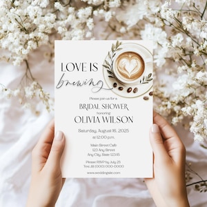 Könnte beinhalten: Eine Brautparty-Einladung mit den Worten "Love is brewing" in eleganter Schrift. Die Einladung zeigt eine Kaffeetasse mit einem herzförmigen Design im Schaum, umgeben von Kaffeebohnen und Grünzeug. Die Einladung wird von Händen gehalten.