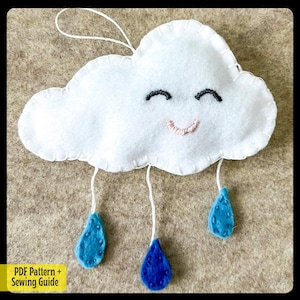 Könnte beinhalten: Eine weiße Filzwolke mit einem lächelnden Gesicht und gestickten Augen. Drei blaue Filztropfen hängen an der Wolke.