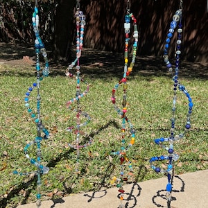 May include: Four colorful beaded wind chimes hanging from a yellow metal bar. Each chime is made of glass beads in various colors, including blue, green, red, yellow, and pink. The chimes are hanging in front of a green grassy lawn.