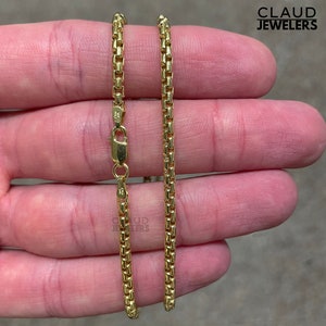 May include: A gold chain necklace with a box chain design. The chain is shown laid out on a hand.
