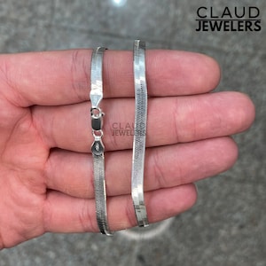 May include: A silver chain necklace with a herringbone pattern. The chain is shown on a hand, with the clasp visible.