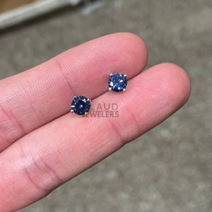 May include: A pair of silver stud earrings with round blue gemstones. The earrings are on a finger.