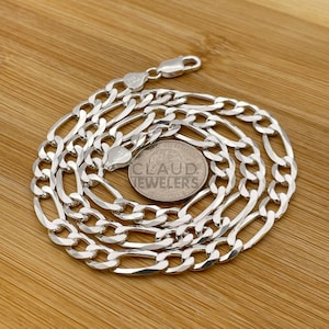 May include: A silver Figaro chain necklace with a flat, oval-shaped link design. The chain is coiled on a wooden surface and a US quarter is placed next to it for size comparison.