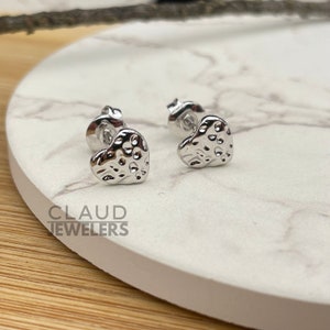May include: A pair of silver heart-shaped stud earrings with a textured surface. The earrings are on a white marble surface.