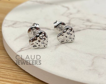 May include: A pair of silver heart-shaped stud earrings with a textured surface. The earrings are on a white marble surface.