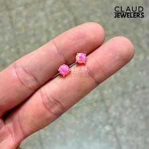May include: A pair of pink opal stud earrings with silver posts. The earrings have a round, polished opal stone with a vibrant pink hue and a subtle yellow and orange color variation. The earrings are held between fingers.