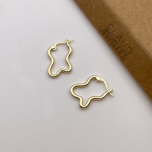 May include: A pair of gold-toned, abstract-shaped hoop earrings. The earrings are made of a smooth, shiny metal and have a slightly irregular shape. The earrings are shown against a white background.