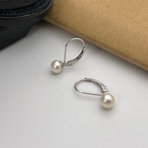 May include: A pair of leverback earrings with white pearls. The earrings have a silver-colored metal finish and a classic design. The pearls are round and have a smooth surface. The earrings are displayed on a white surface.