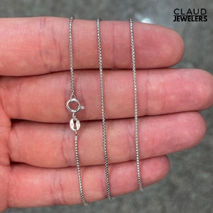 May include: A silver chain necklace with a box clasp. The chain is thin and delicate, and it is shown lying on a hand.