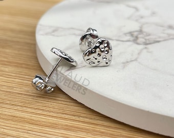 May include: A pair of silver heart-shaped stud earrings with a textured surface. The earrings are on a white marble surface.