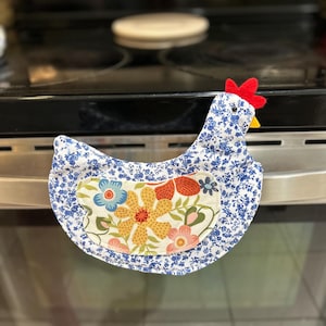 Puede incluir: Un guante de cocina de tela con forma de pollo. El guante presenta un estampado floral azul con una cresta roja y un pico amarillo. El centro tiene un diseño floral con flores amarillas, naranjas y rosas.
