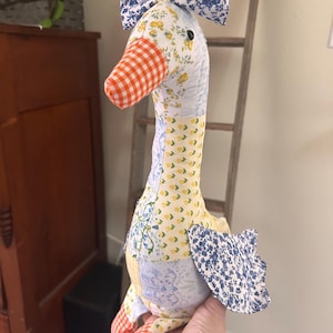May include: A handmade goose doll with a patchwork design. The goose features a bow, an orange gingham beak, and wings. The body is a mix of floral and patterned fabrics in blue, yellow, and white. The goose is being held by a person.