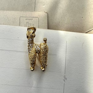 Puede incluir: Un broche de color dorado en forma de caniche, decorado con brillantes transparentes.