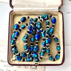 May include: A long necklace made of oval-shaped iridescent blue and green glass beads with black and clear glass beads interspersed throughout. The necklace is displayed in a red velvet jewelry box.