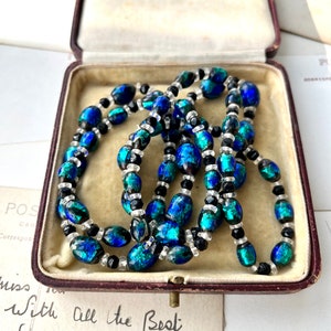 May include: A vintage necklace made of iridescent blue and green glass beads with black and clear glass beads. The necklace is in a red velvet jewelry box.