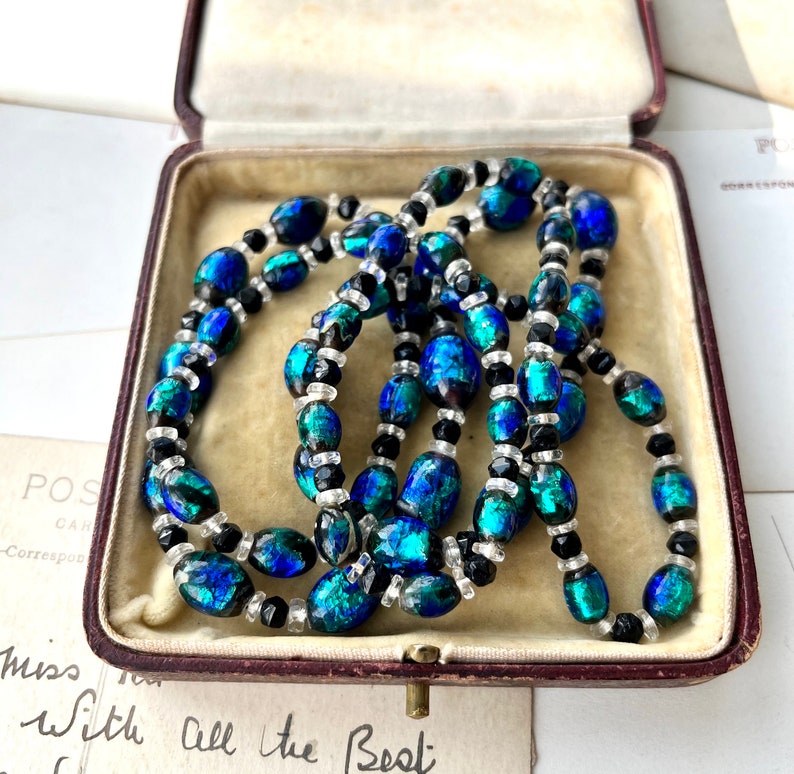 May include: A vintage necklace made of iridescent blue and green glass beads with black and clear glass beads. The necklace is in a red velvet jewelry box.