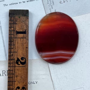 May include: Oval-shaped, polished gemstone with bands of red, orange, and white. A vintage wooden ruler with black markings is positioned next to the stone. The ruler shows measurements up to 3 inches. The word "CARD" is visible.