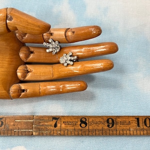 May include: Two silver tone brooches with clear rhinestones. The brooches are on a wooden hand, and a ruler is in the background.