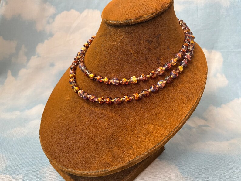 May include: A double strand necklace made of iridescent glass beads with gold flecks. The beads are round and have a textured surface. The necklace is displayed on a brown velvet mannequin.