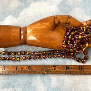 May include: A long strand of iridescent glass beads in shades of brown, gold, and purple. The beads are approximately 1/2 inch in diameter and are strung on a clear thread. The beads are displayed on a wooden hand.