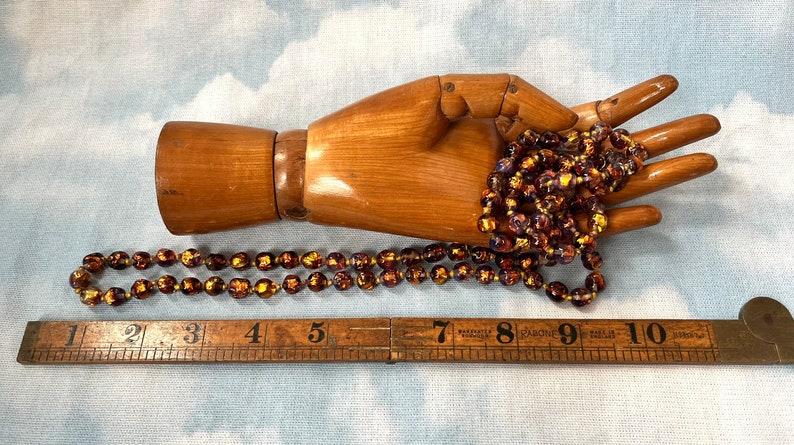 May include: A long strand of iridescent glass beads in shades of brown, gold, and purple. The beads are approximately 1/2 inch in diameter and are strung on a clear thread. The beads are displayed on a wooden hand.