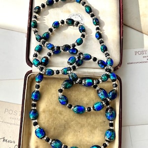 May include: A vintage necklace made of iridescent blue and green glass beads with black and clear glass accents. The necklace is displayed in a velvet-lined jewelry box.