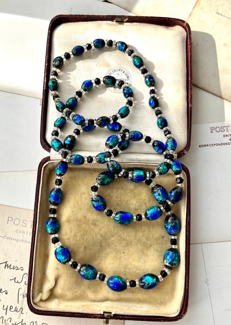 May include: A vintage necklace made of iridescent blue and green glass beads with black and clear glass accents. The necklace is displayed in a velvet-lined jewelry box.