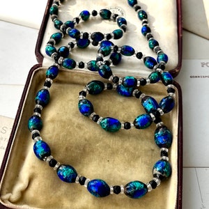 May include: A vintage necklace made of oval-shaped iridescent blue and green glass beads interspersed with black and clear round beads. The necklace is displayed in a brown velvet-lined box.