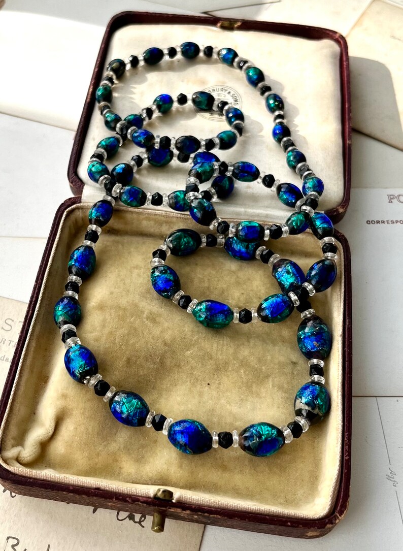 May include: A vintage necklace made of oval-shaped iridescent blue and green glass beads interspersed with black and clear round beads. The necklace is displayed in a brown velvet-lined box.
