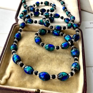 May include: A long necklace and a shorter bracelet made of oval-shaped iridescent blue and green glass beads with black and clear spacer beads.